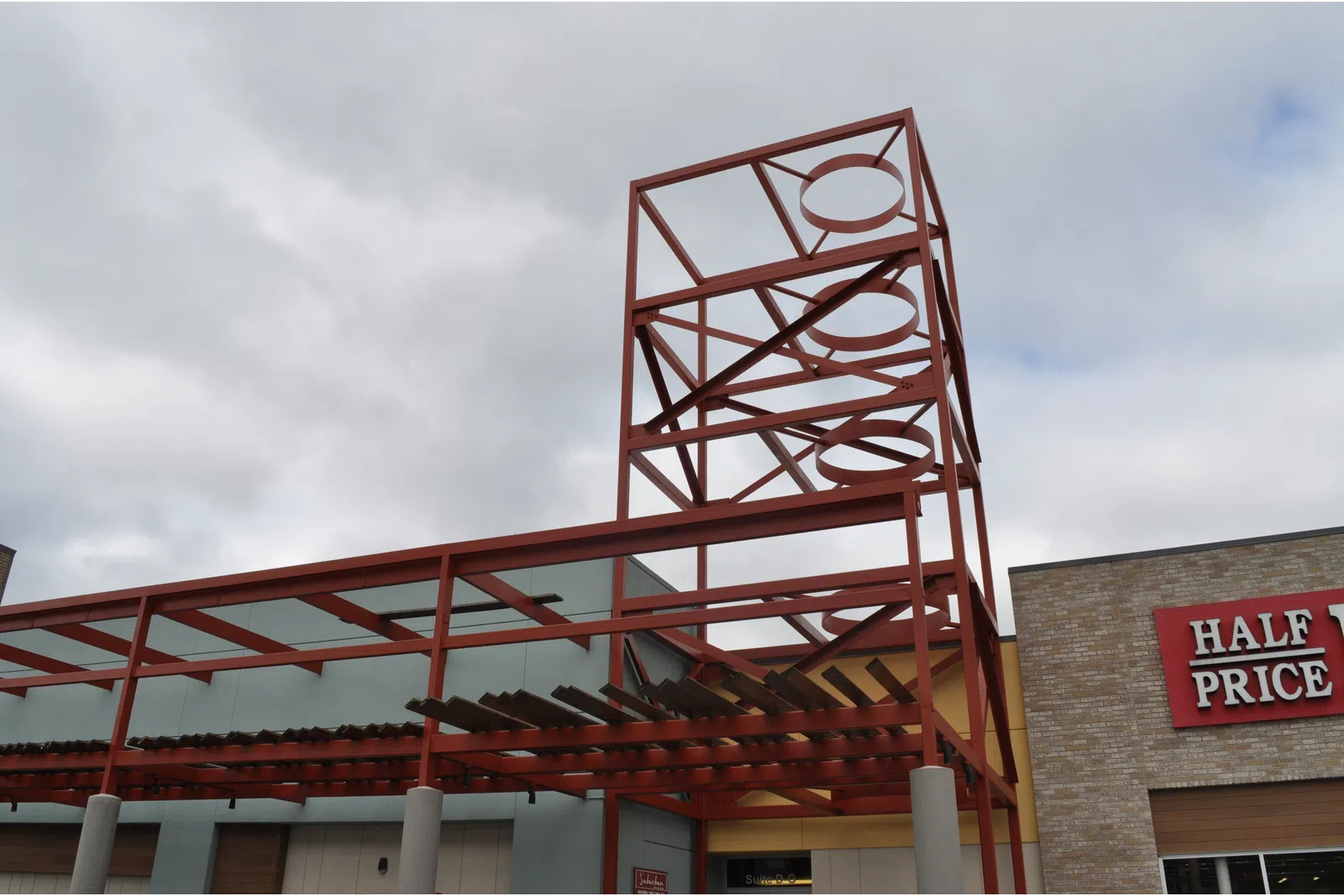 Red metal framework structure with circular cutouts above a building entrance under cloudy sky.