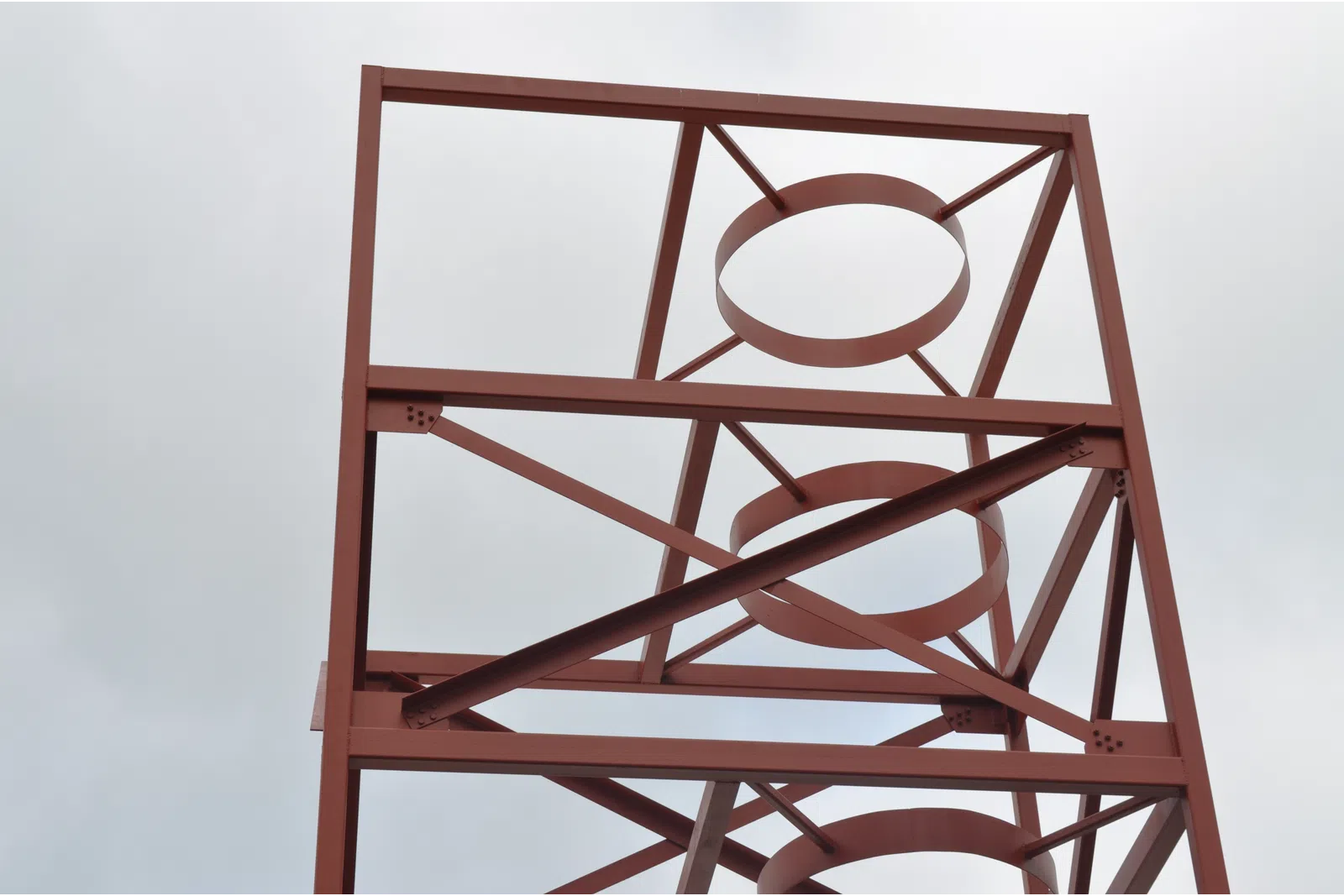 Rust-colored metal framework with circular and diagonal supports against a cloudy sky.