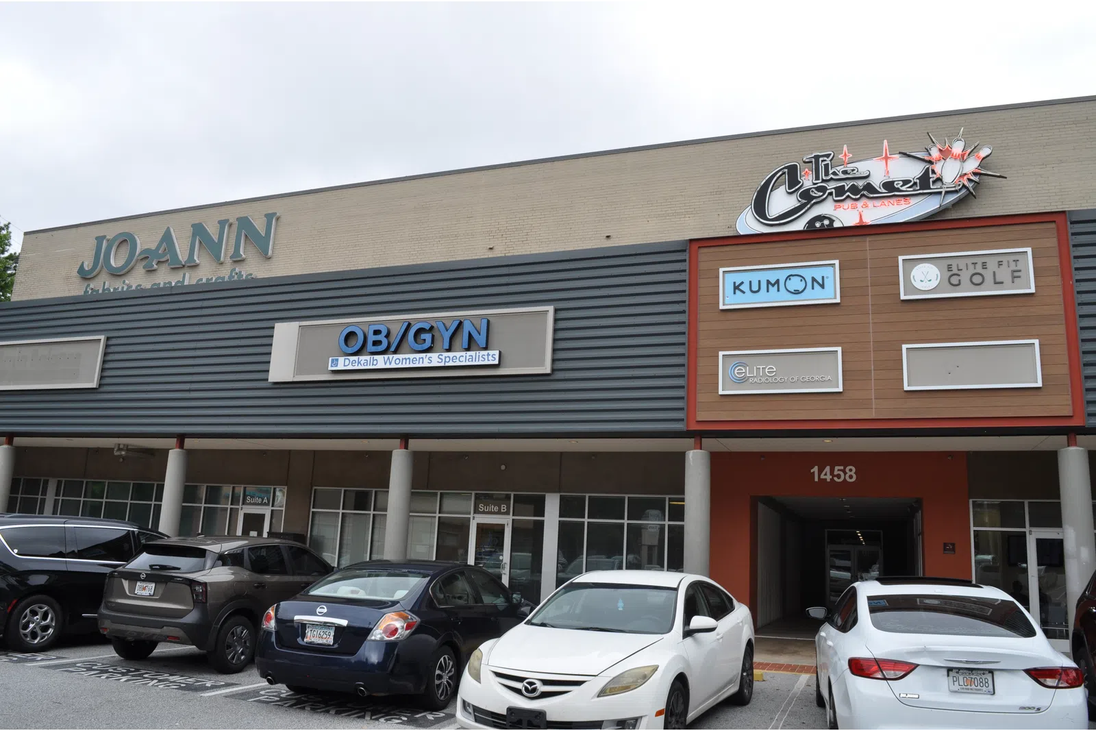 Shopping plaza exterior with storefronts including JOANN, OB/GYN, Kumon, and parking lot with cars in front.