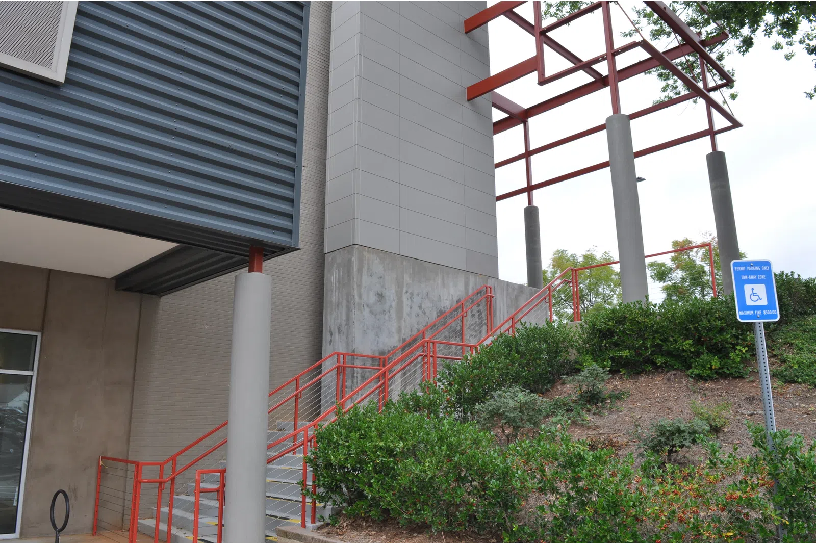 Outdoor staircase with red railings next to modern building and green shrubbery under metal canopy.