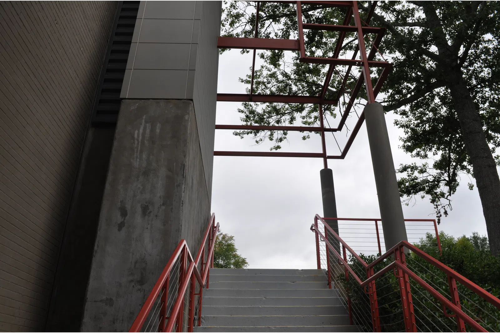 Concrete stairs with red railings lead up to a metal frame structure surrounded by trees against a cloudy sky.