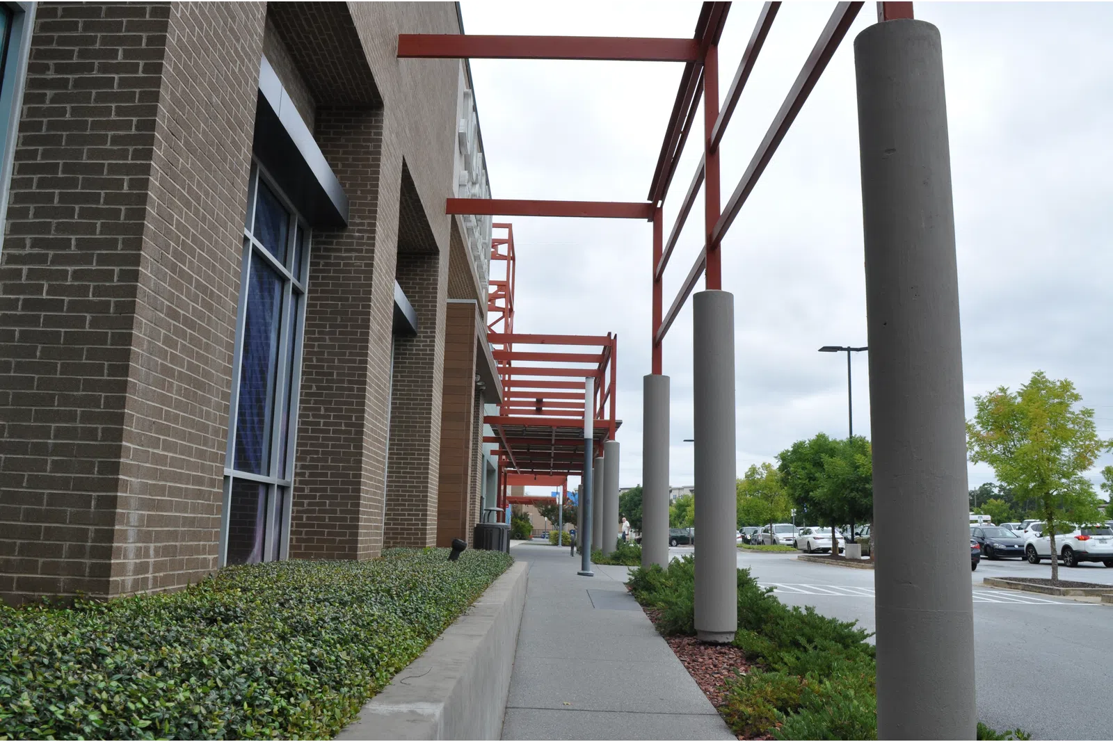 Sidewalk beside a brick building with red steel beams and concrete pillars under an overcast sky near a parking lot.