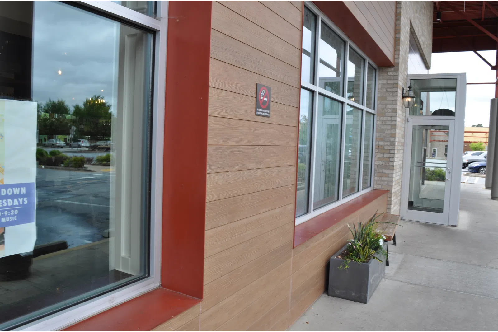 Modern restaurant exterior with large windows, tan wood paneling, potted plants, and sidewalk seating area.