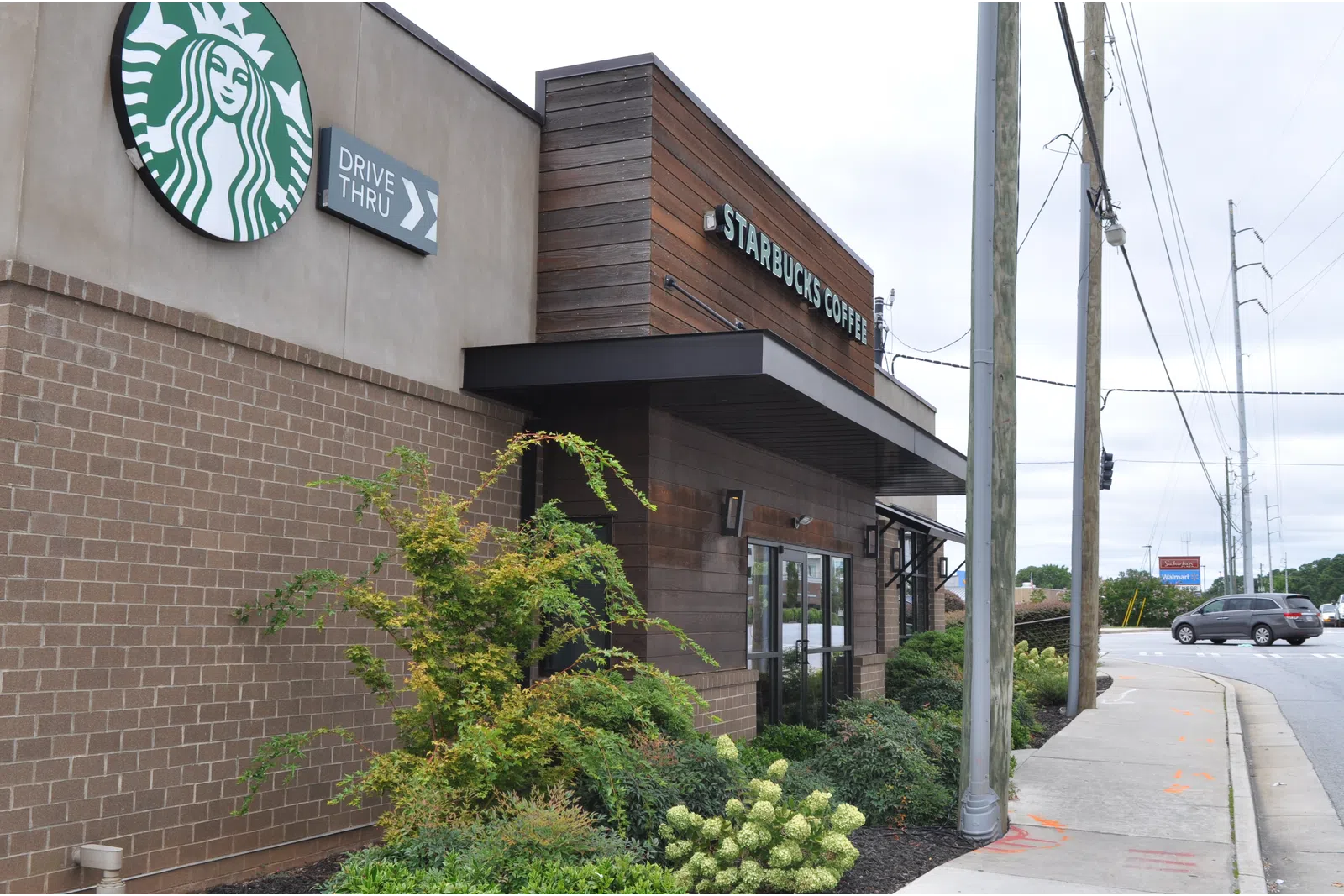 Starbucks coffee shop exterior with drive-thru sign and landscaped greenery by busy street corner.
