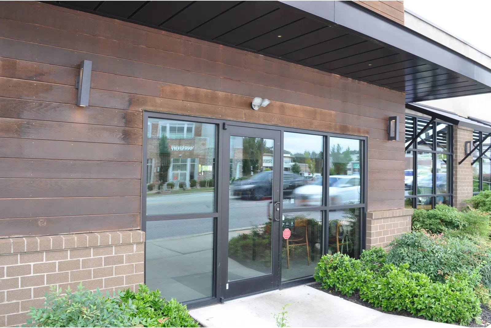 Glass double doors with black frames at storefront reflecting street and cars, surrounded by wood and brick exterior with greenery.