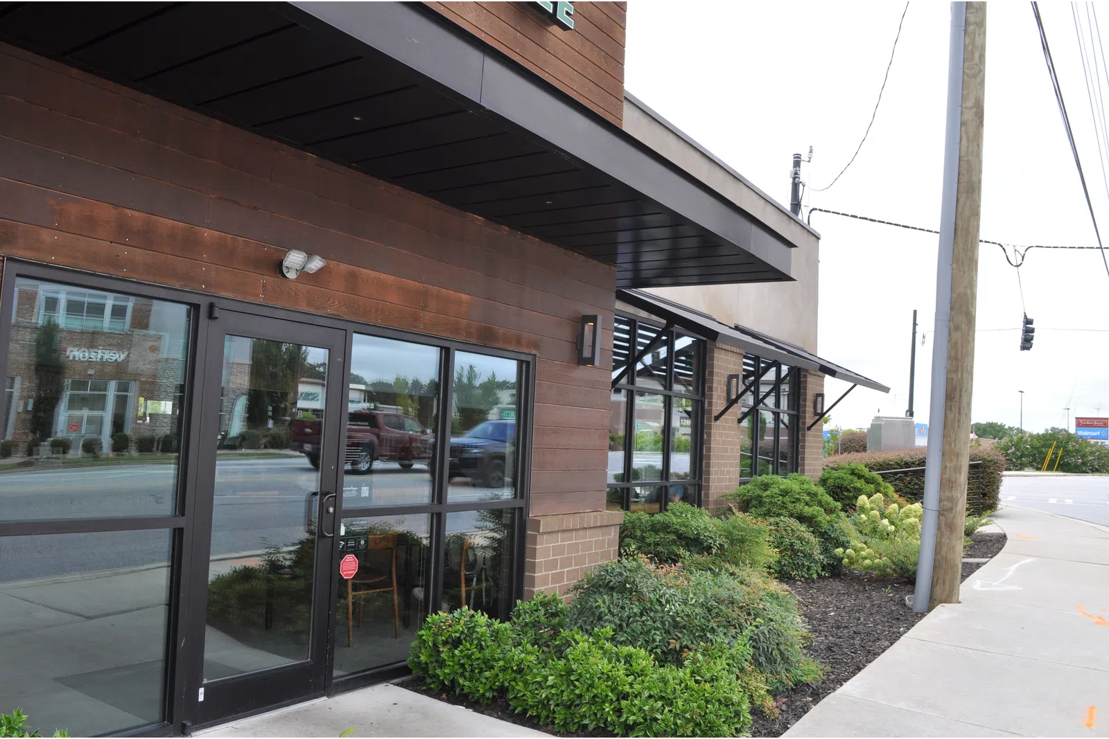 Modern brown wooden storefront with glass doors and windows, surrounded by green shrubs along a sidewalk.