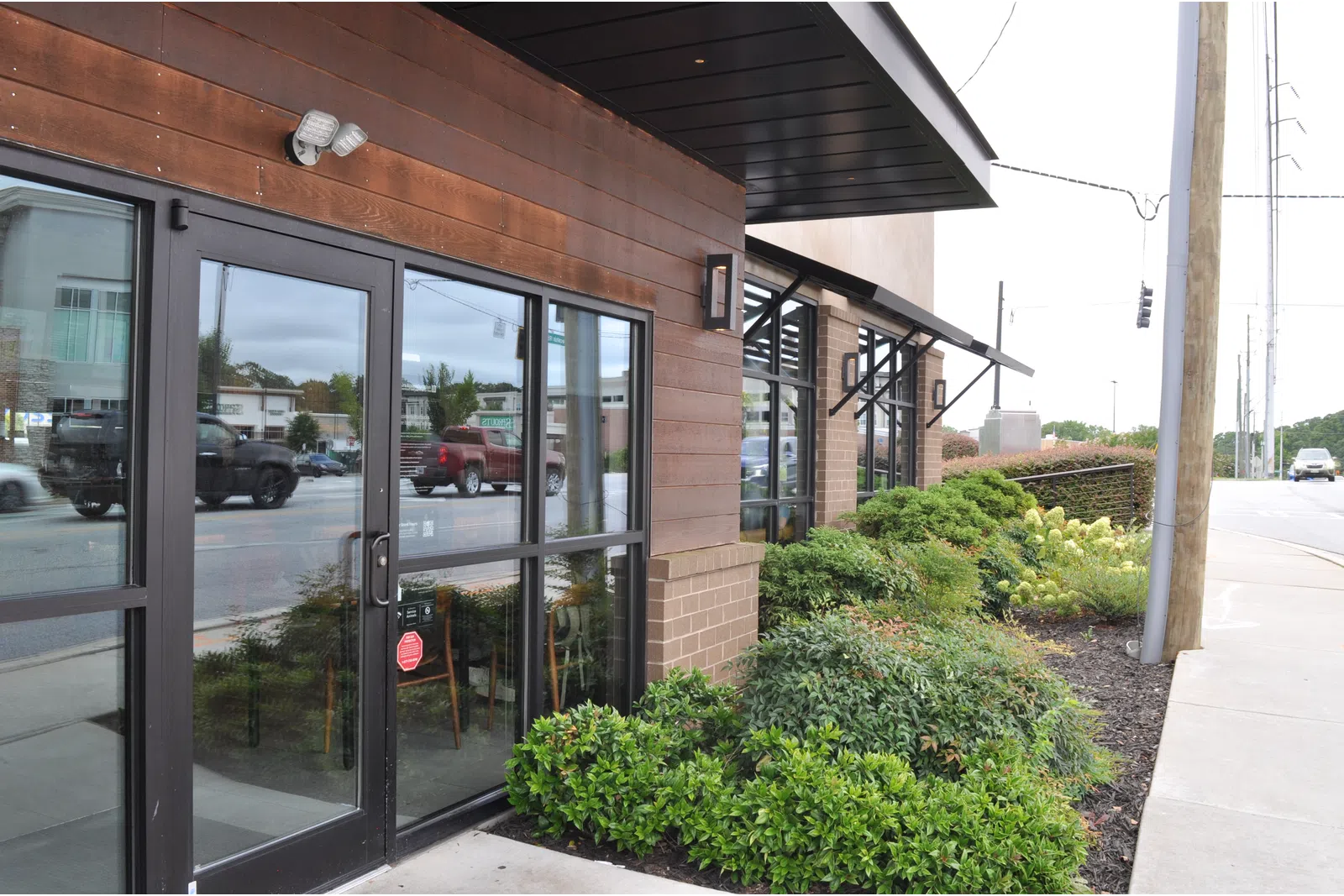 Commercial building entrance with glass doors and landscaped greenery along the sidewalk on a busy street.