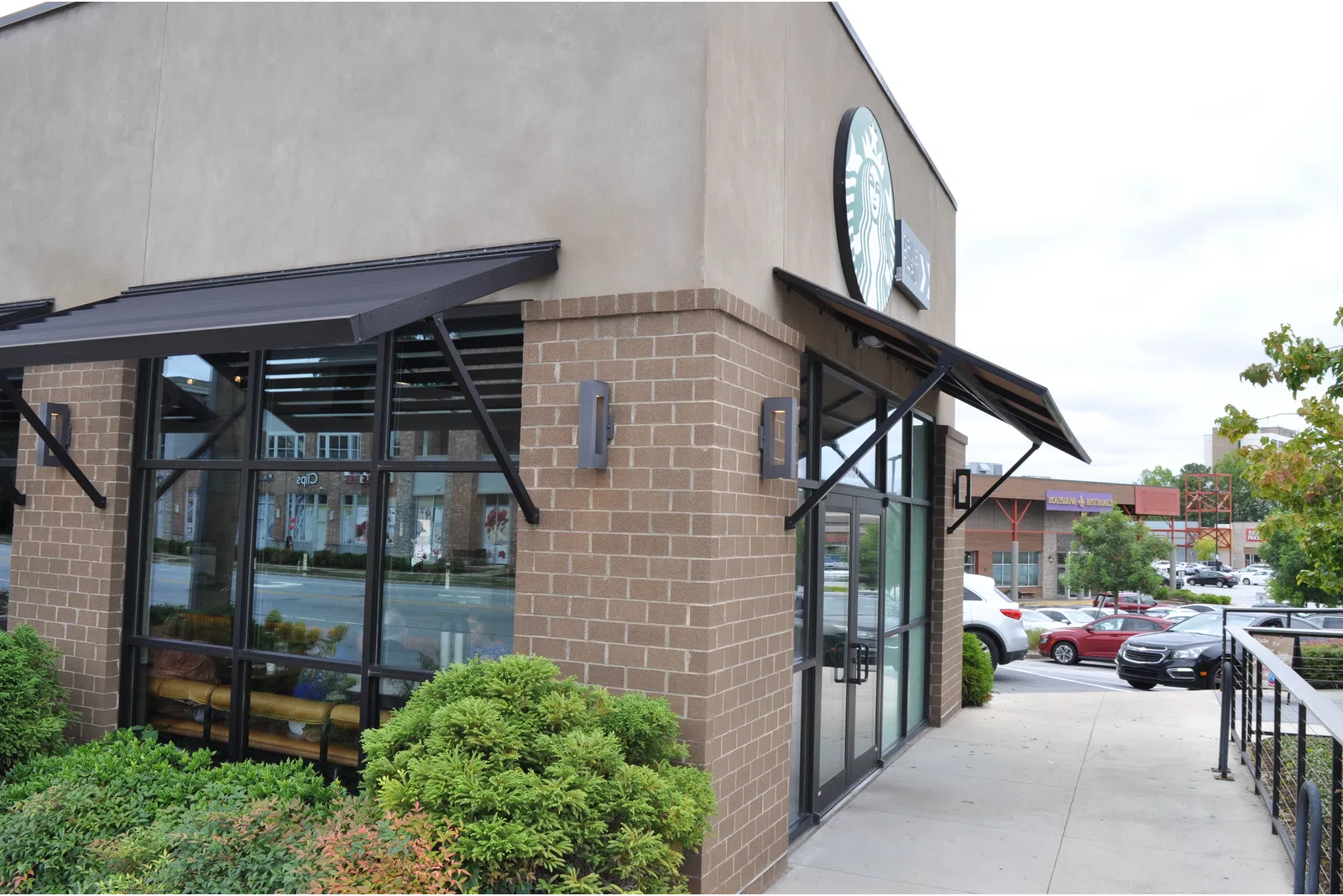 Exterior view of a Starbucks coffee shop with brick walls, large windows, and outdoor greenery near a parking lot.