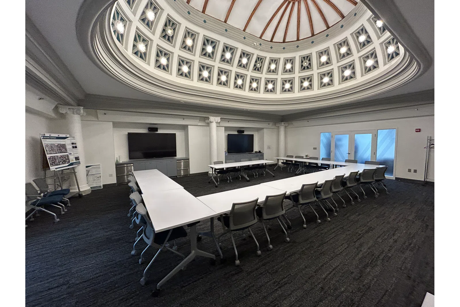 Modern conference room with U-shaped white tables, rolling chairs, and a decorative domed ceiling with lights.