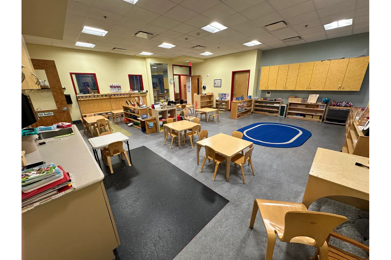 Spacious preschool classroom with small tables, chairs, shelves, and a blue carpet area for group activities.