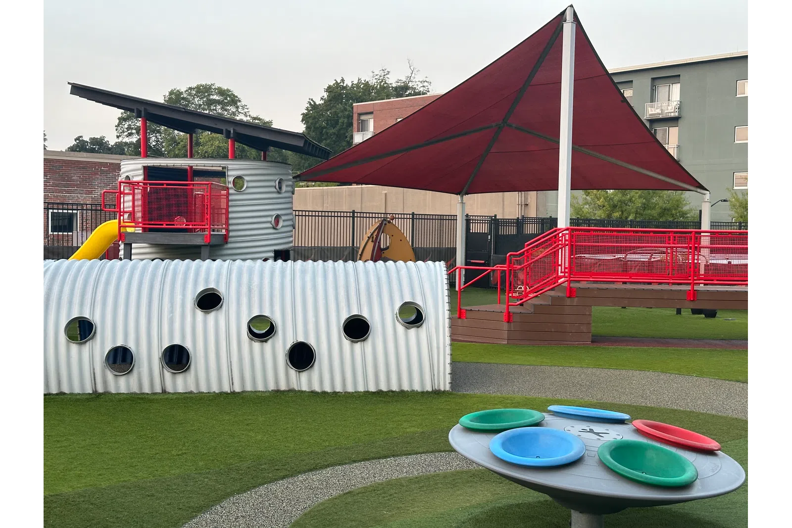 Modern playground with a large red shade canopy, corrugated tunnel, colorful spinning play equipment, and a slide.