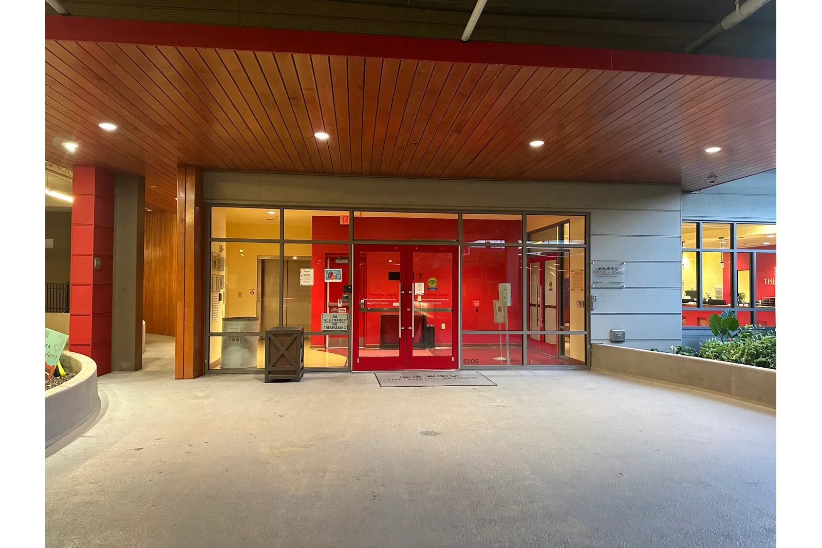 Entrance to a modern building with red doors, glass panels, wooden ceiling, and concrete floor at dusk