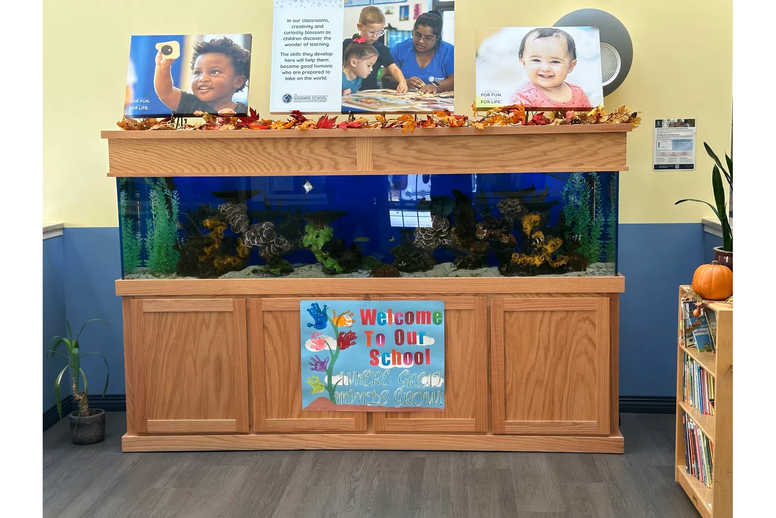 Large aquarium with decorative plants in a school lobby featuring welcome signs and children's photos above it.