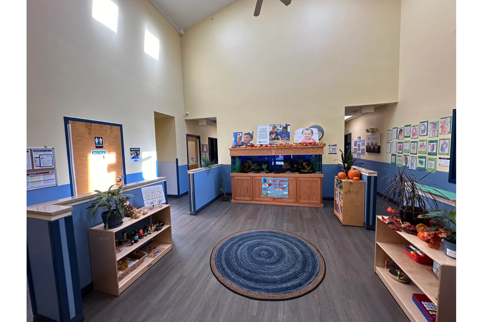 Bright, spacious daycare lobby with blue circular rug, fish tank, shelves with toys, and children's artwork on walls.