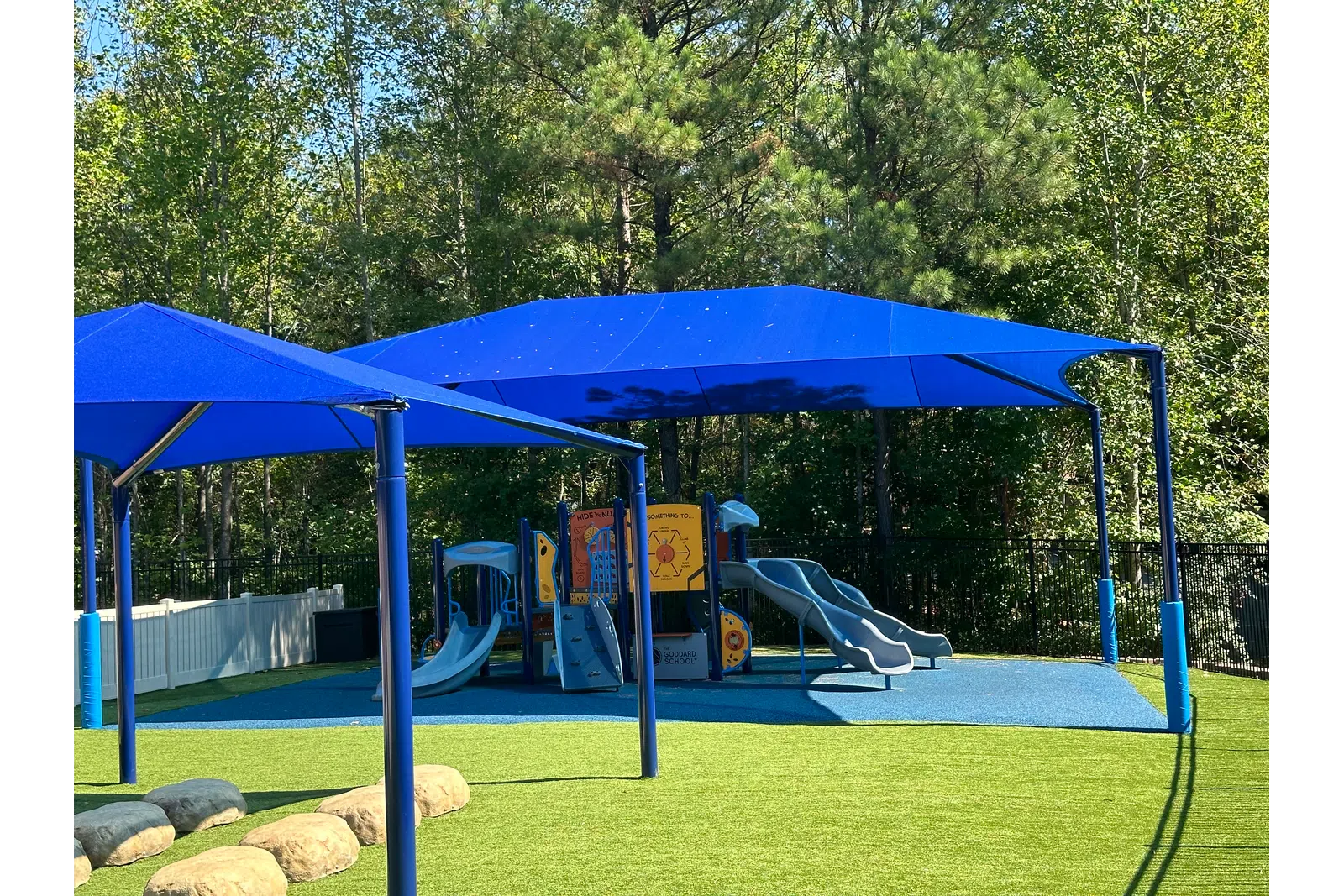 Outdoor playground with blue slides, climbing structures, and blue shade canopies surrounded by greenery.