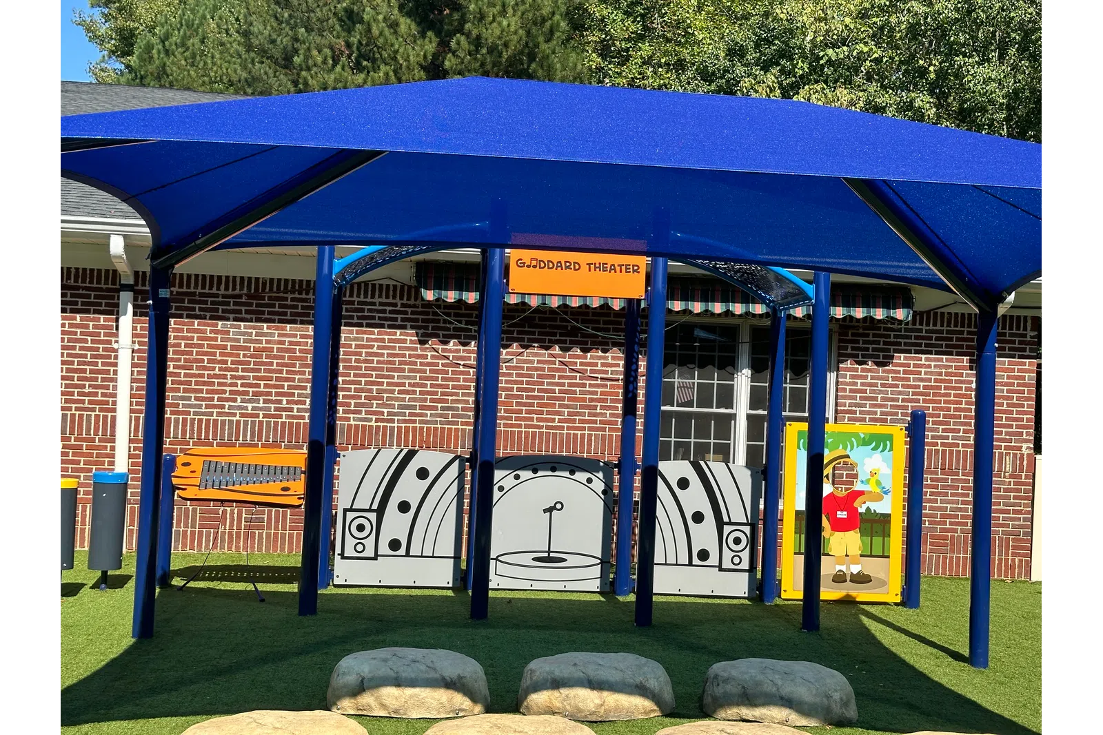 Outdoor children's play theater area with blue canopy, stage backdrop, and artificial grass seating resembling rocks.