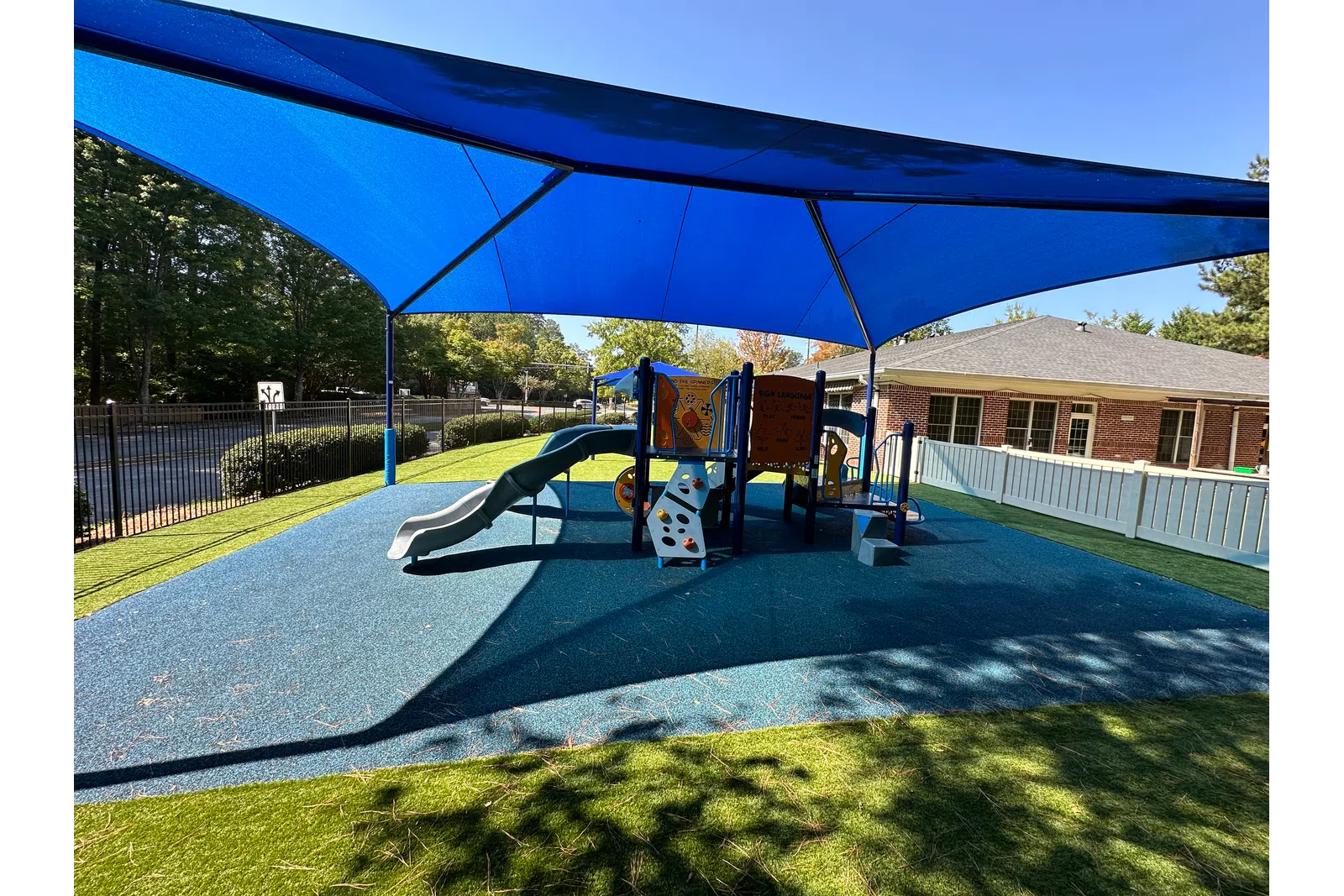 Outdoor playground with slides and climbing structures shaded by large blue canopy on sunny day.