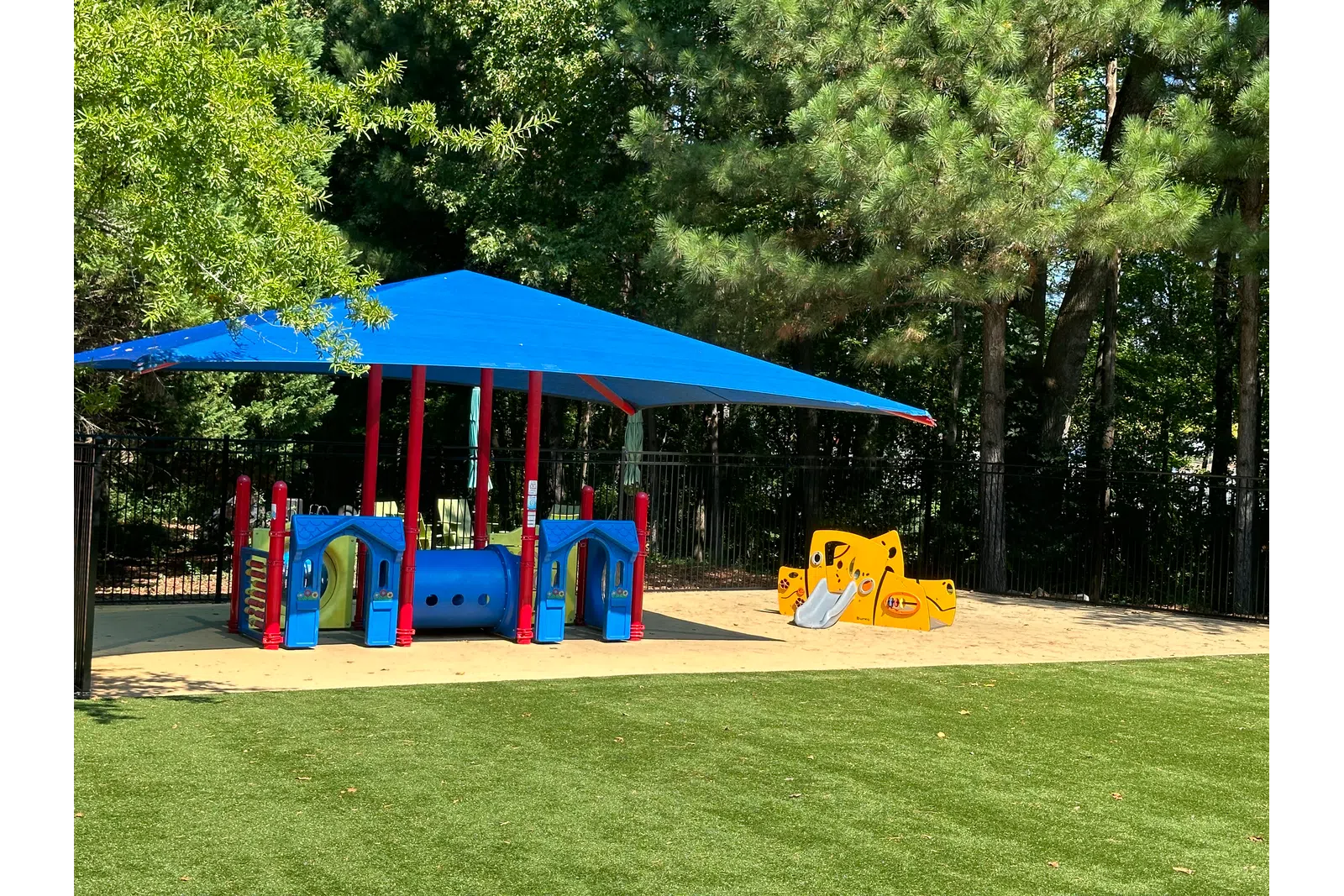 Children's playground with blue shade canopy, colorful play structures, slides, and artificial grass surrounded by trees.