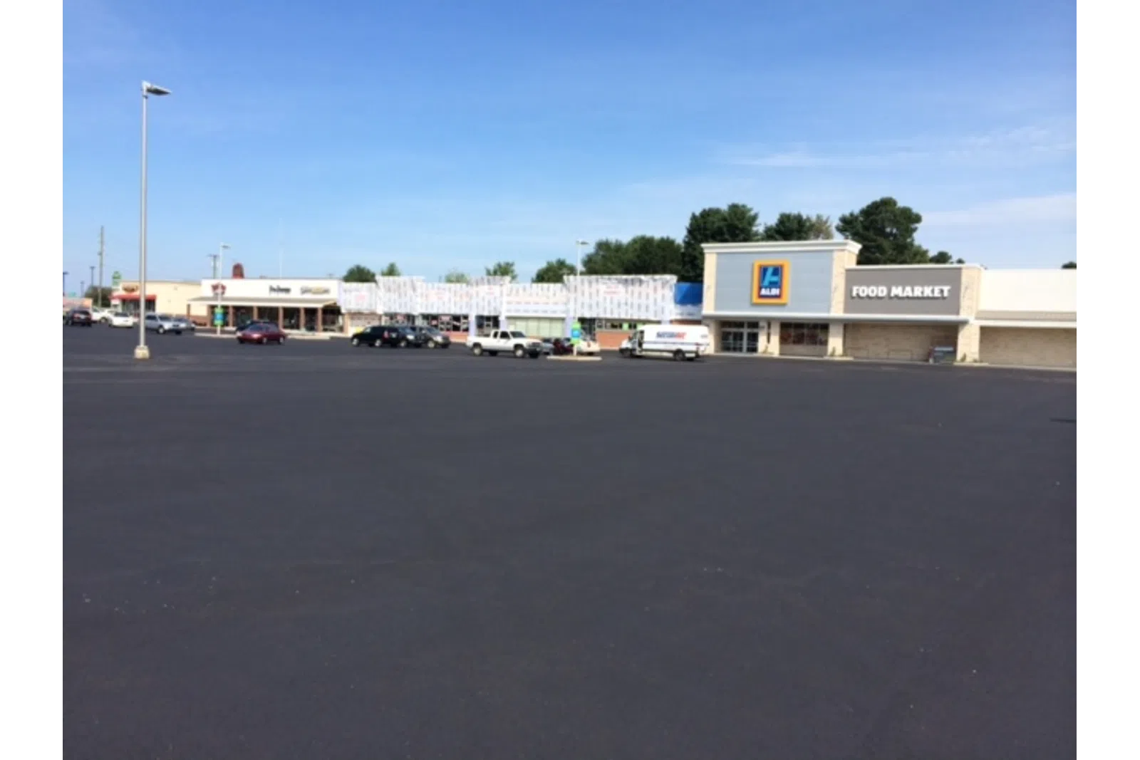 Wide empty parking lot in front of a strip mall featuring Aldi Food Market and other stores under clear blue sky