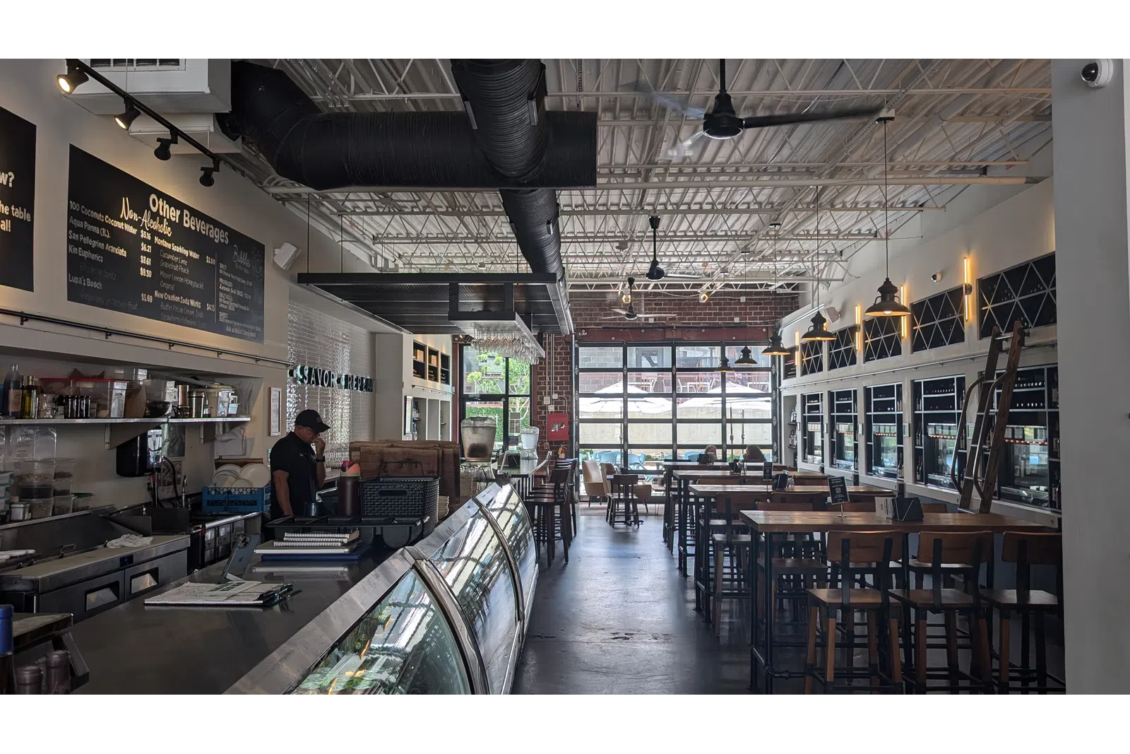 Interior of a modern cafe with high ceilings, wooden tables, industrial decor, and a glass display counter.