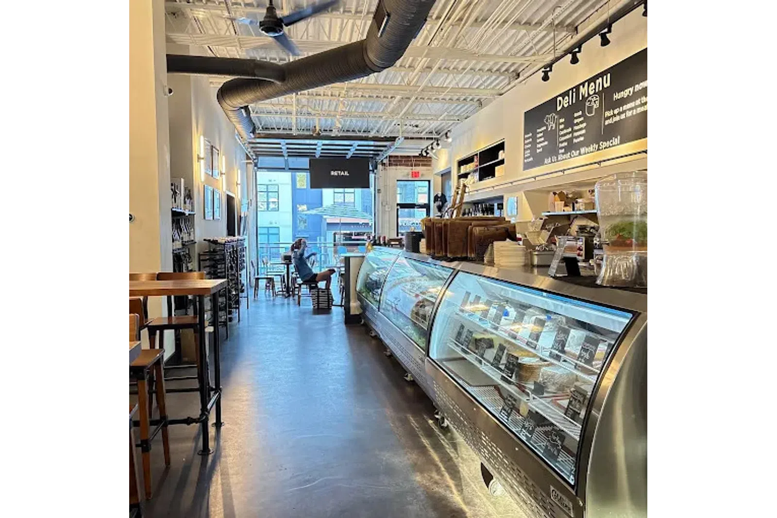 Bright modern deli interior with glass display cases, seating area, and a person sitting near the entrance.