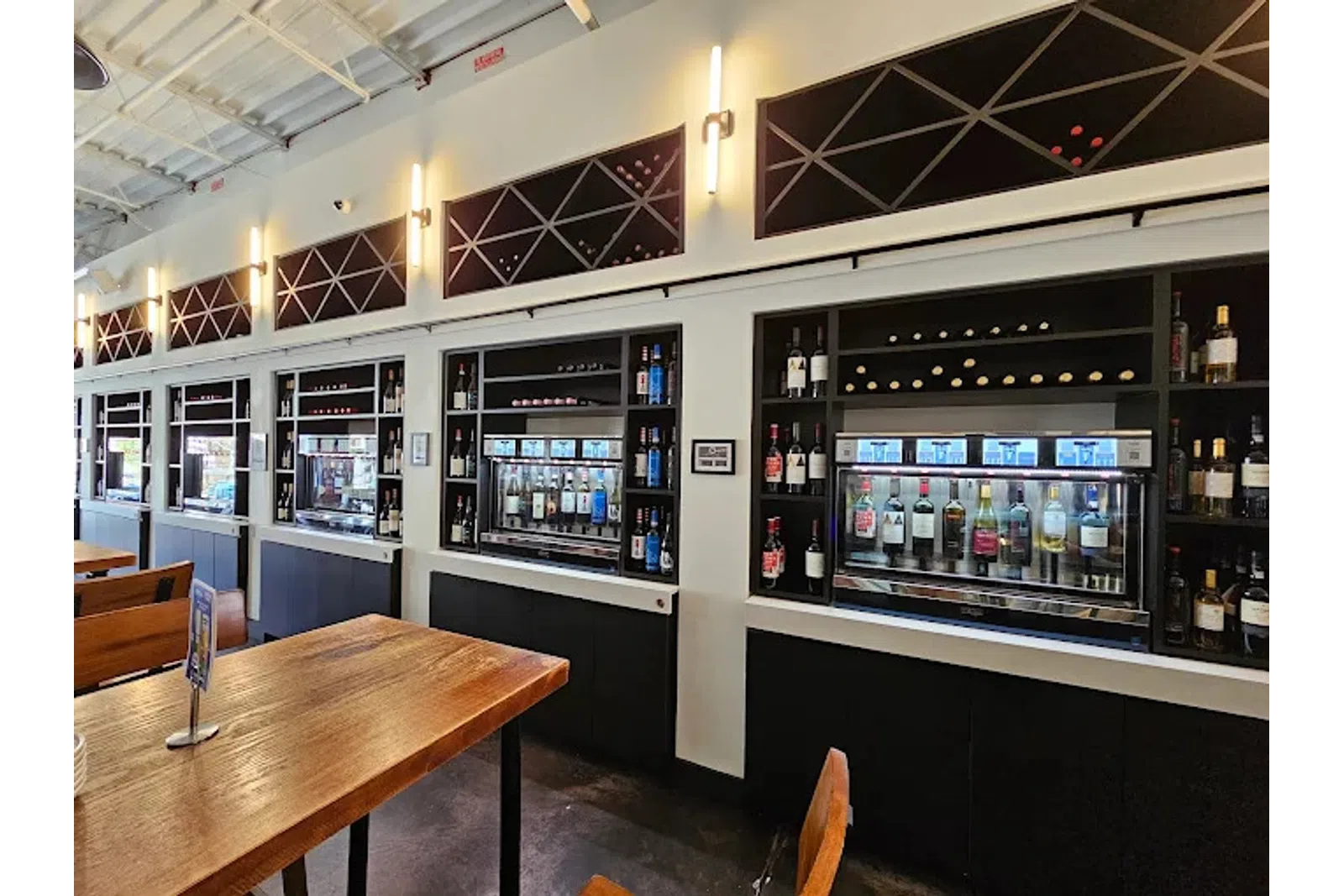 Modern wine tasting room with wooden tables and automated wine dispensers against white walls.