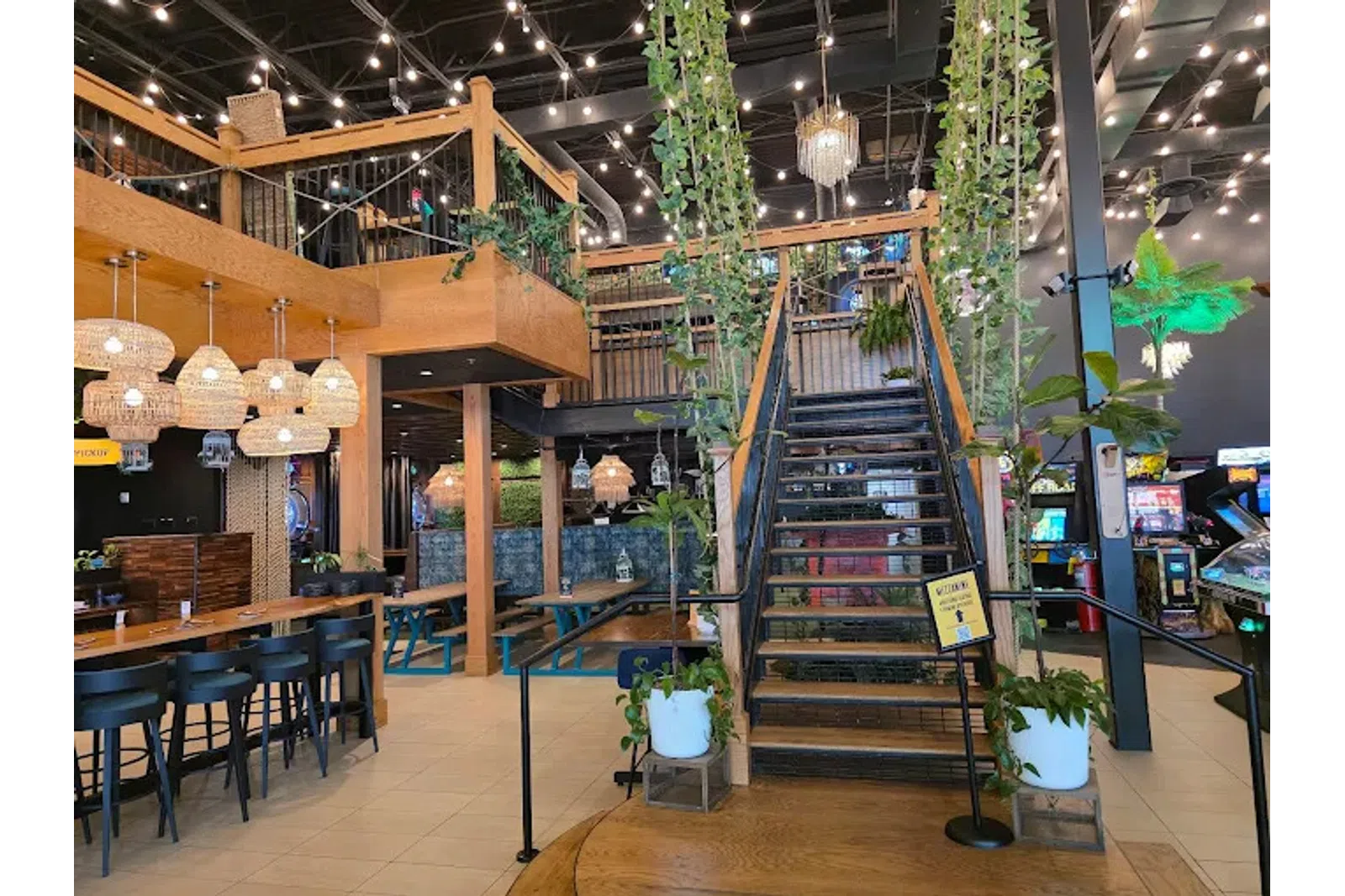 Modern indoor restaurant with wooden tables, hanging lights, plants, staircase, and arcade games in the background