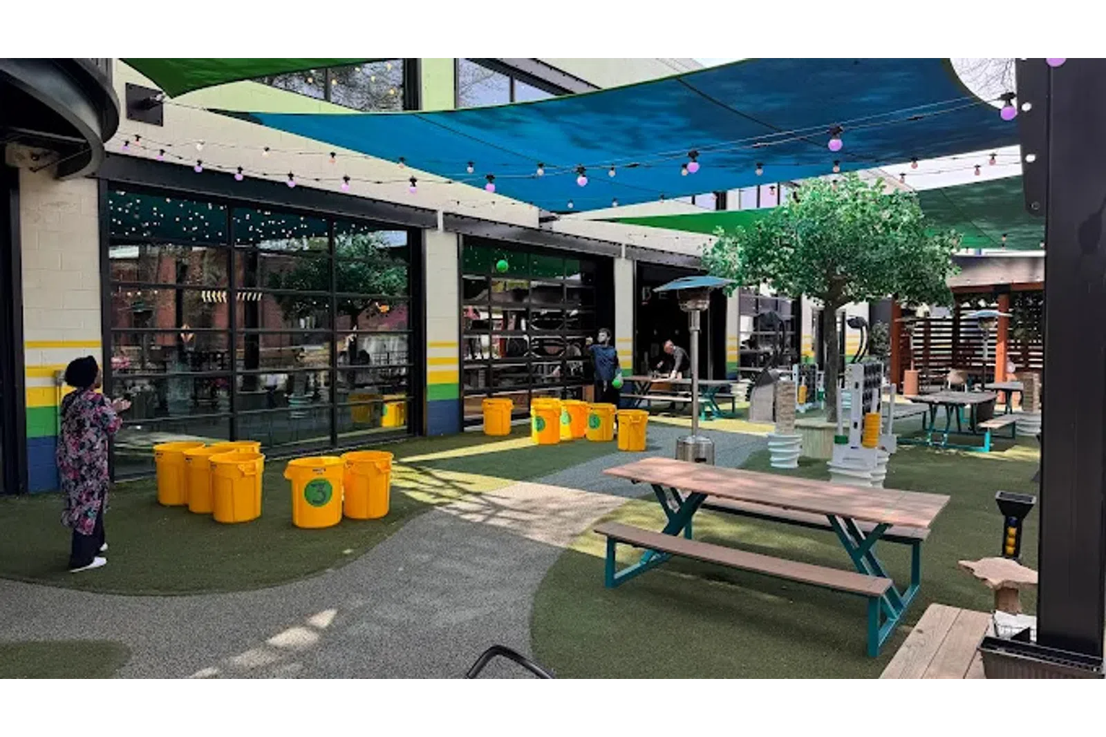 Outdoor seating area with picnic tables, yellow recycling bins, shade sails, and string lights in a commercial space.
