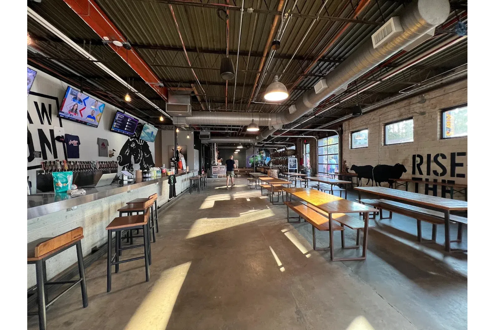 Spacious brewery tasting room with bar stools, wooden tables, industrial ceiling, and wall art of cows and text.