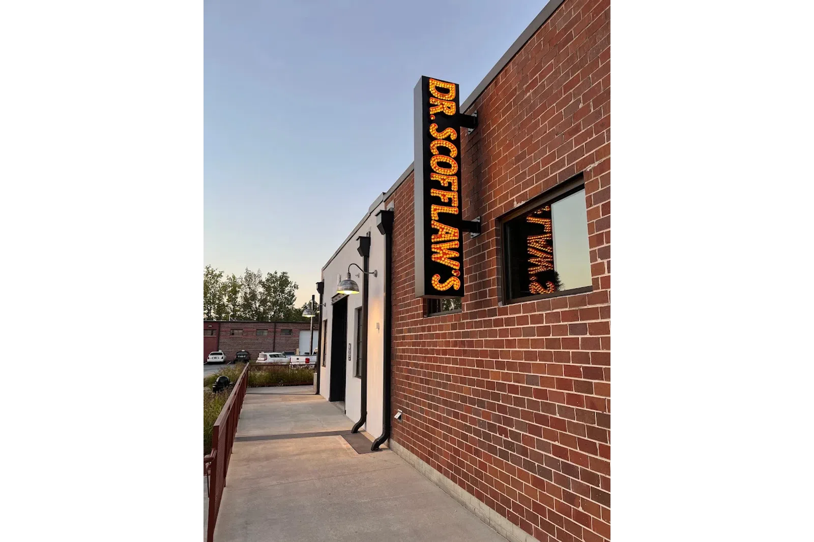 Lit vertical sign reading Dr. Scoflaws mounted on a brick building facade with sidewalk and street lamps.