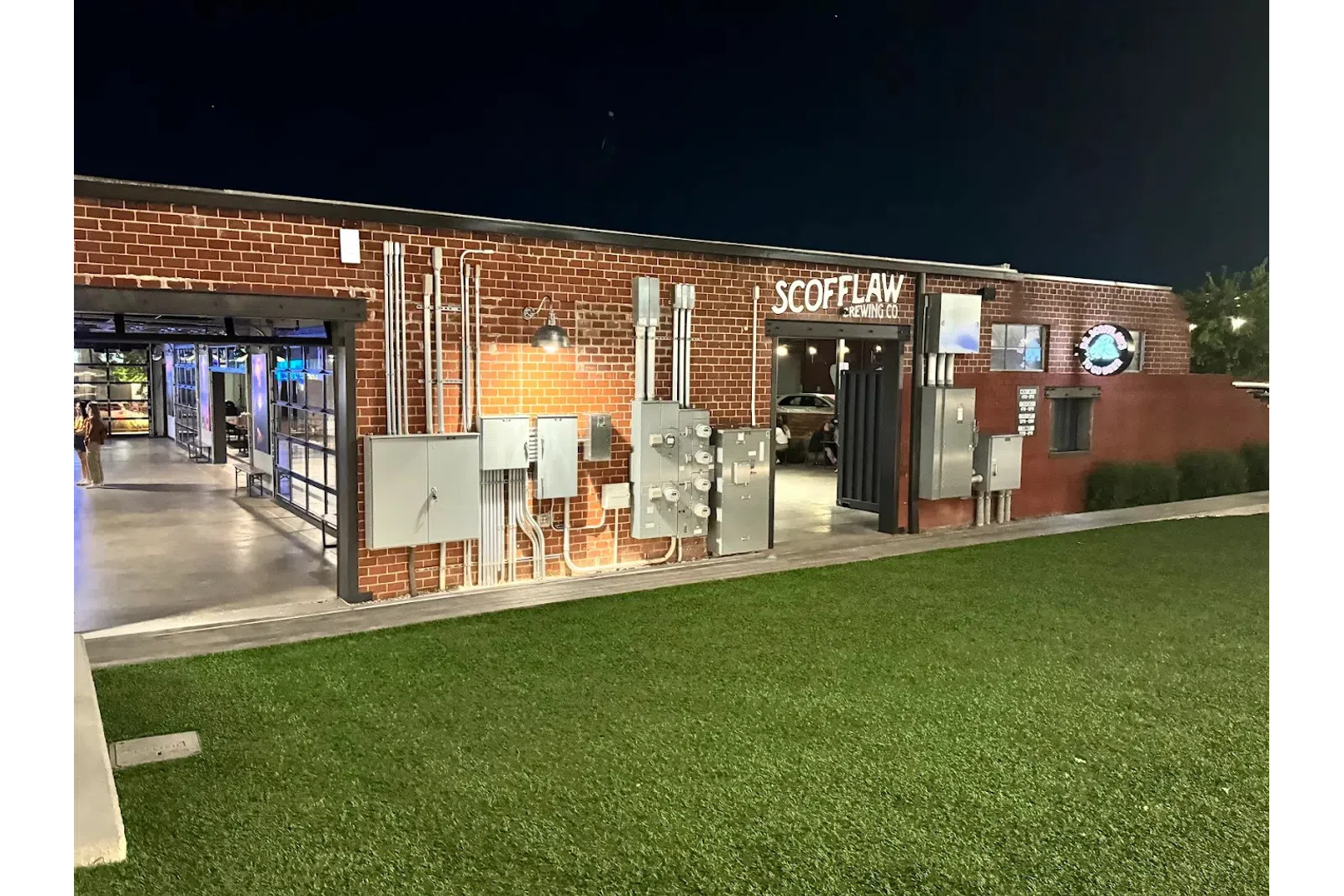 Night view of Scofflaw Brewing Co building with exposed brick, electrical panels, and green lawn in front.