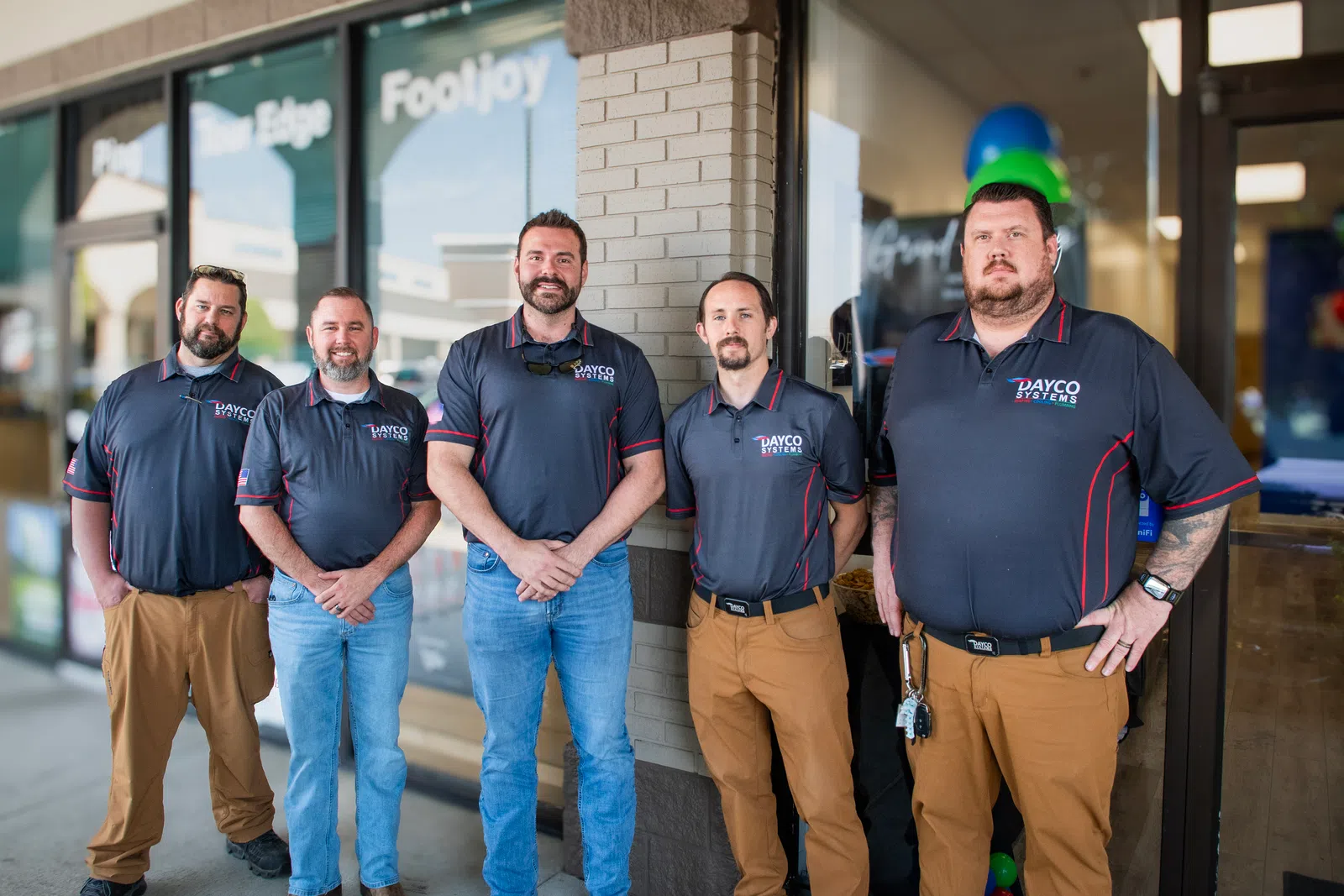 Five men in matching Dayco Systems shirts and khaki pants standing outside a storefront in a row.