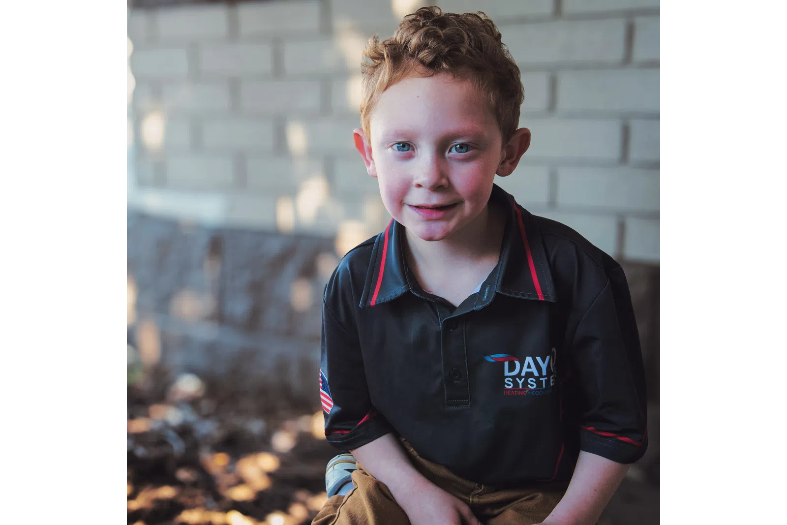 Young boy with curly red hair wearing a black Dayco Systems shirt outdoors near a brick wall.