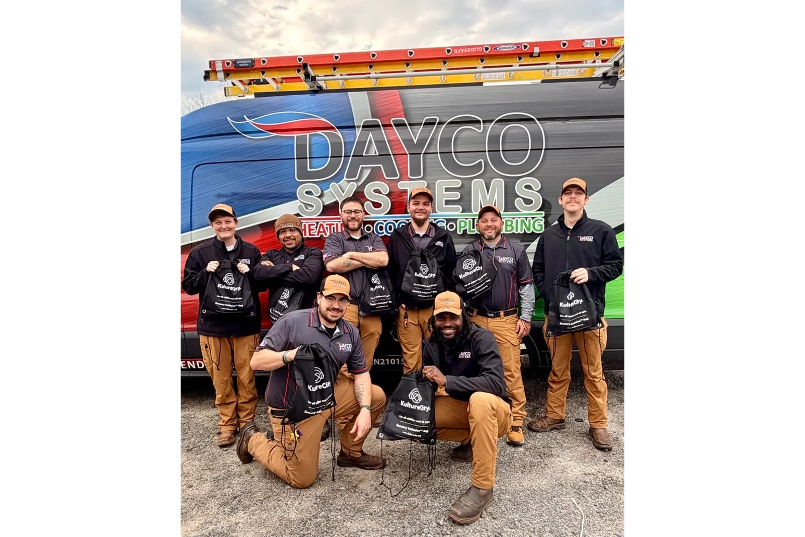 Dayco Systems team of technicians posing with branded bags in front of a company van with ladder on top.