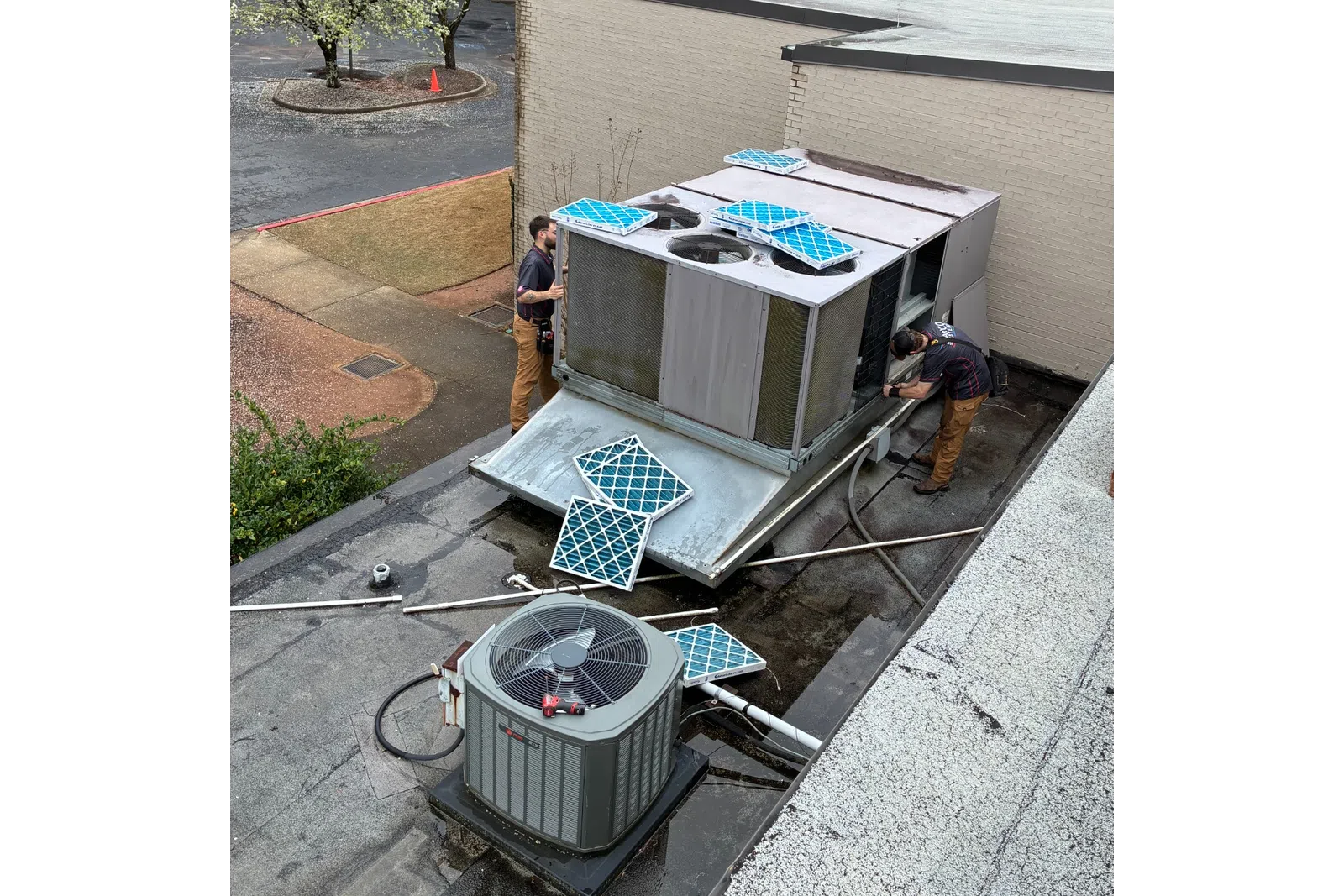 Technicians installing or repairing a rooftop HVAC unit with new blue filters on a flat roof beside a building.