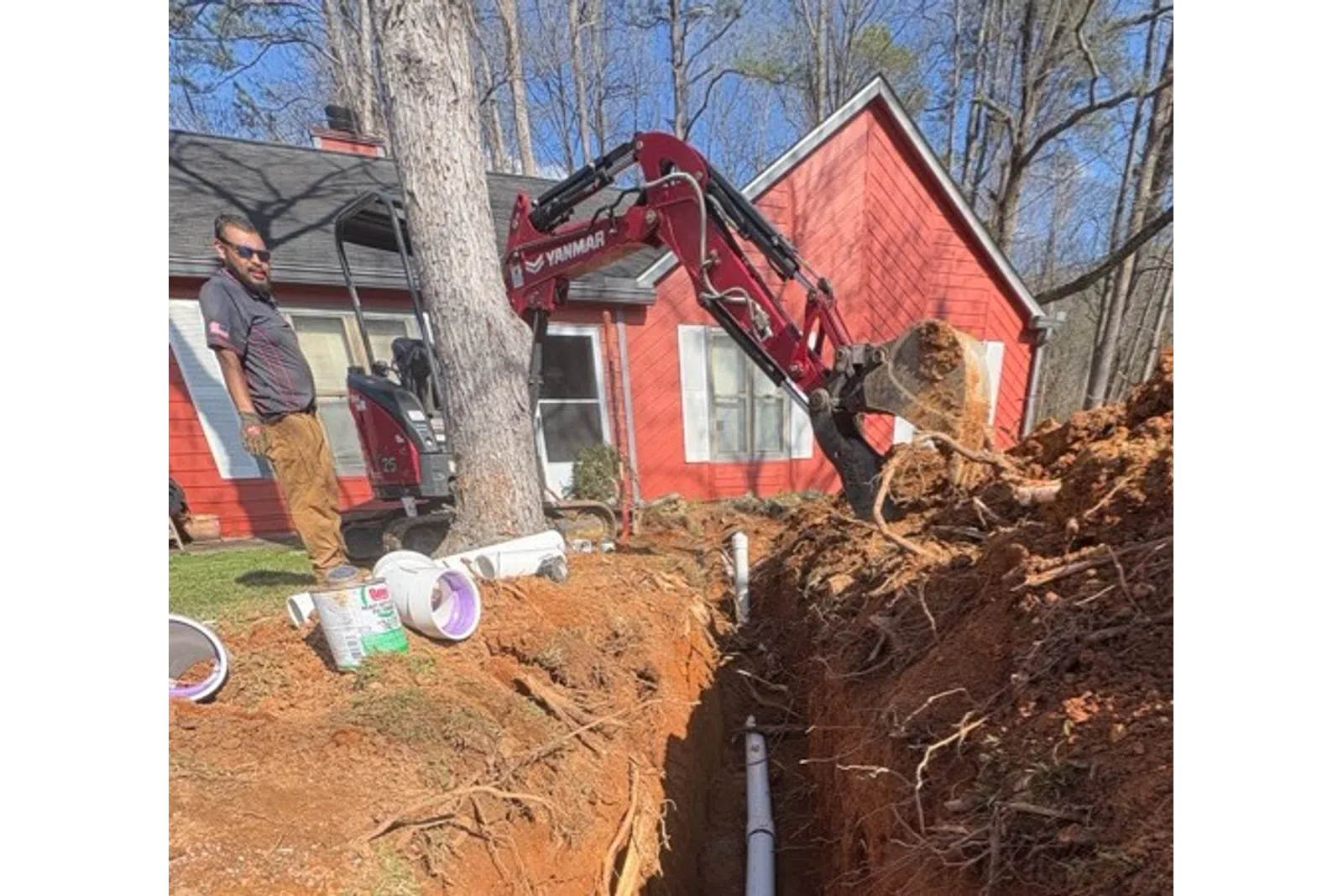 Excavator digging trench for white PVC pipe installation near red house in a wooded area.