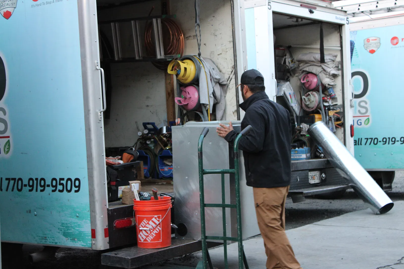 Worker unloading metal duct and tools from a service truck using a hand truck on a city street.