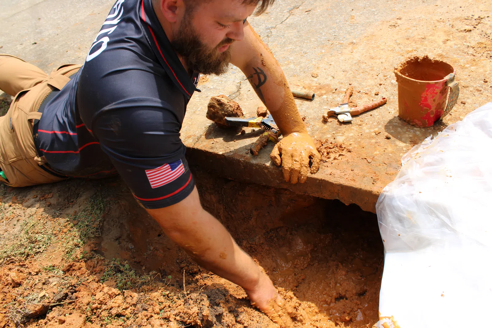Archaeologist excavating clay soil near a concrete slab using hands and tools outdoors on a sunny day.