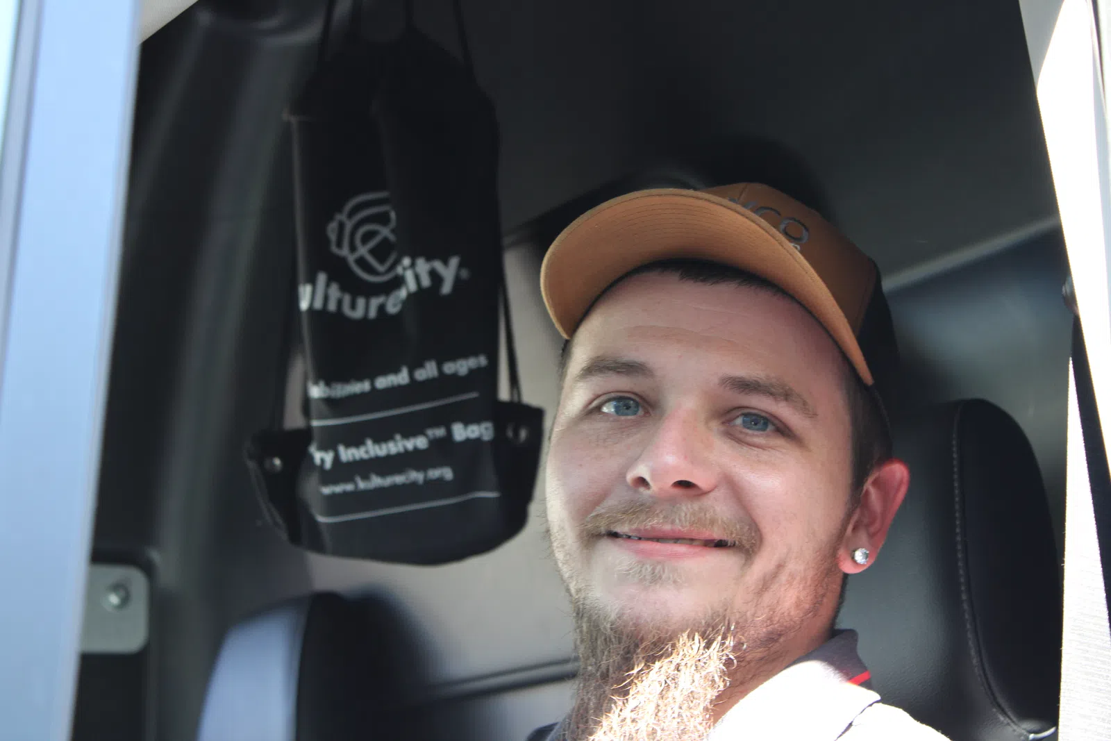 Smiling man with beard and cap sitting inside a vehicle near a hanging black CultureCity bag.