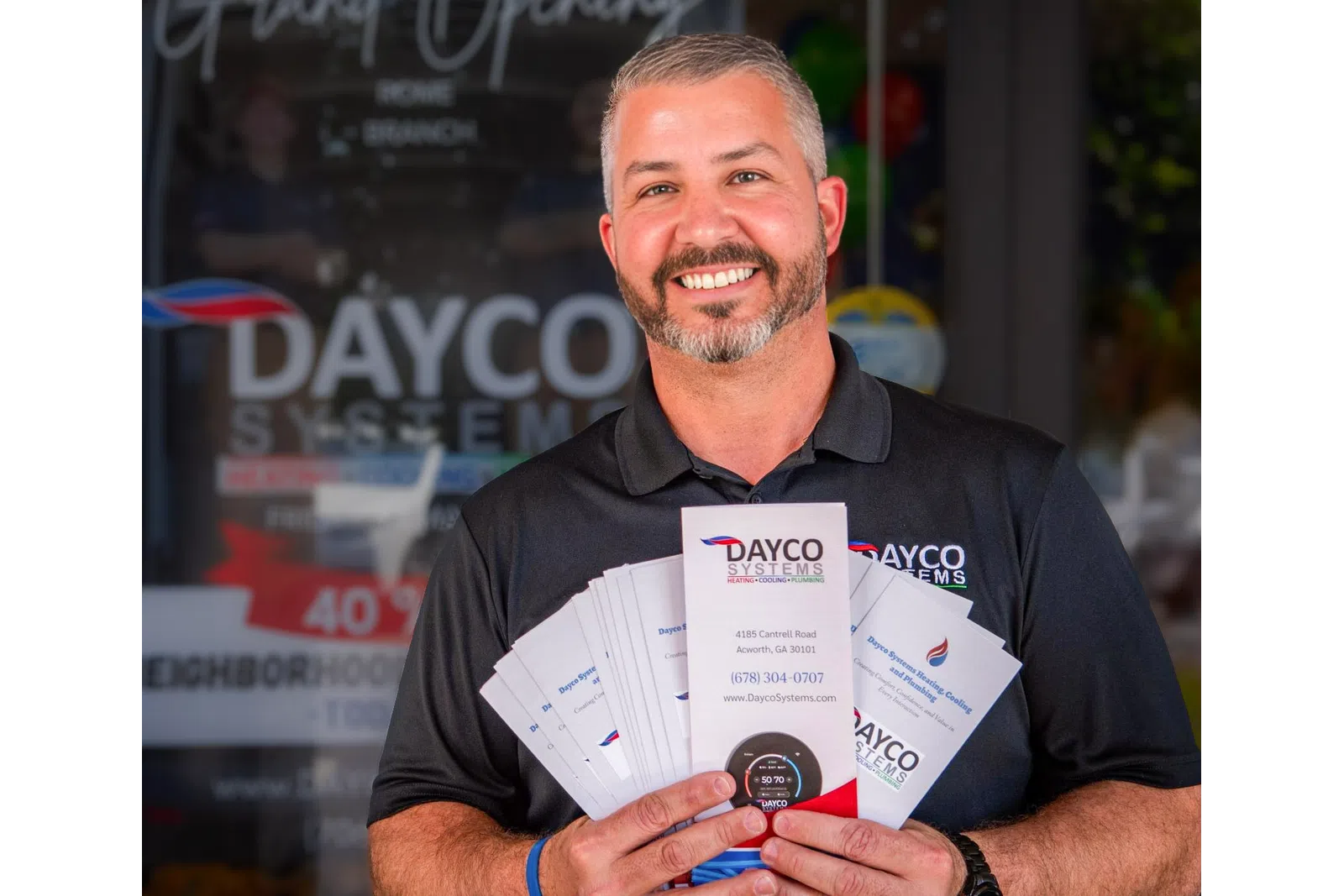 Smiling man in black Dayco Systems shirt holding fan of company brochures outside business entrance.