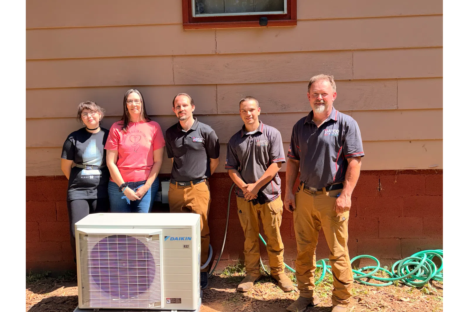 Five people standing outdoors behind a Daikin air conditioning unit next to a red and beige building wall.