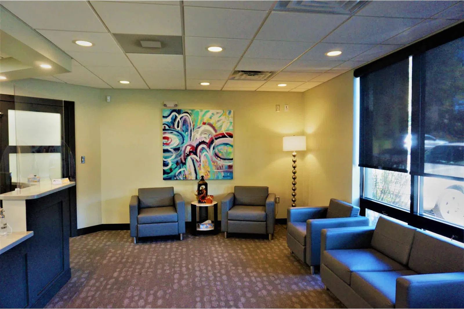 Modern waiting room with gray chairs, colorful abstract painting, floor lamp, and large windows with dark blinds.
