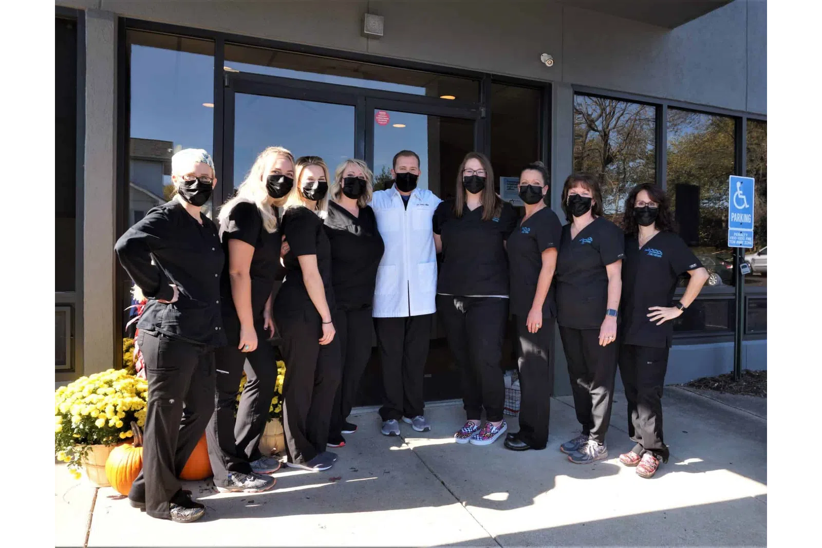 Group of healthcare workers wearing black scrubs and masks standing outside a clinic entrance with autumn decorations.