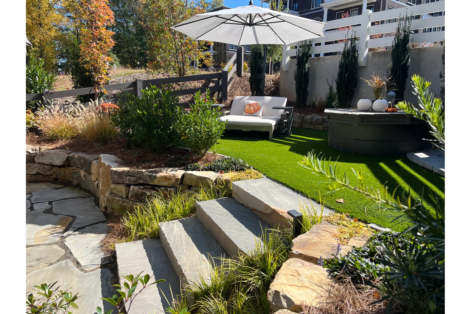 Cozy backyard patio with stone steps, white umbrella, cushioned seating, green grass, and autumn plants.