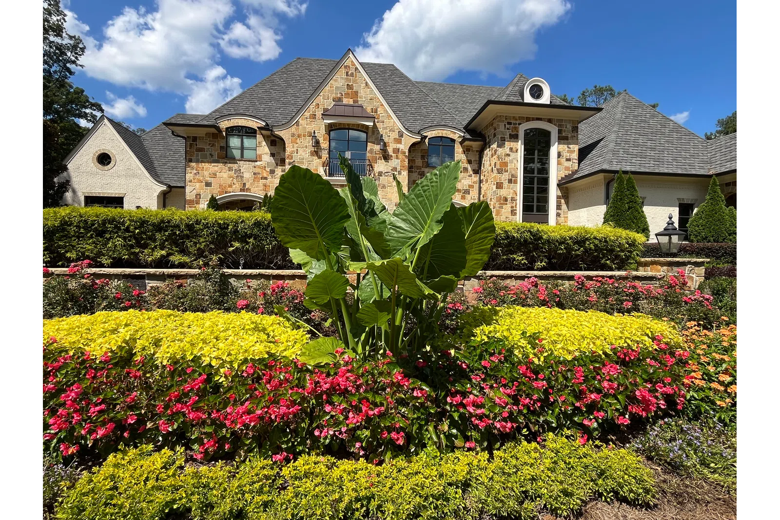 Elegant stone house with manicured garden featuring bright green plants and vibrant pink flowers under blue sky.