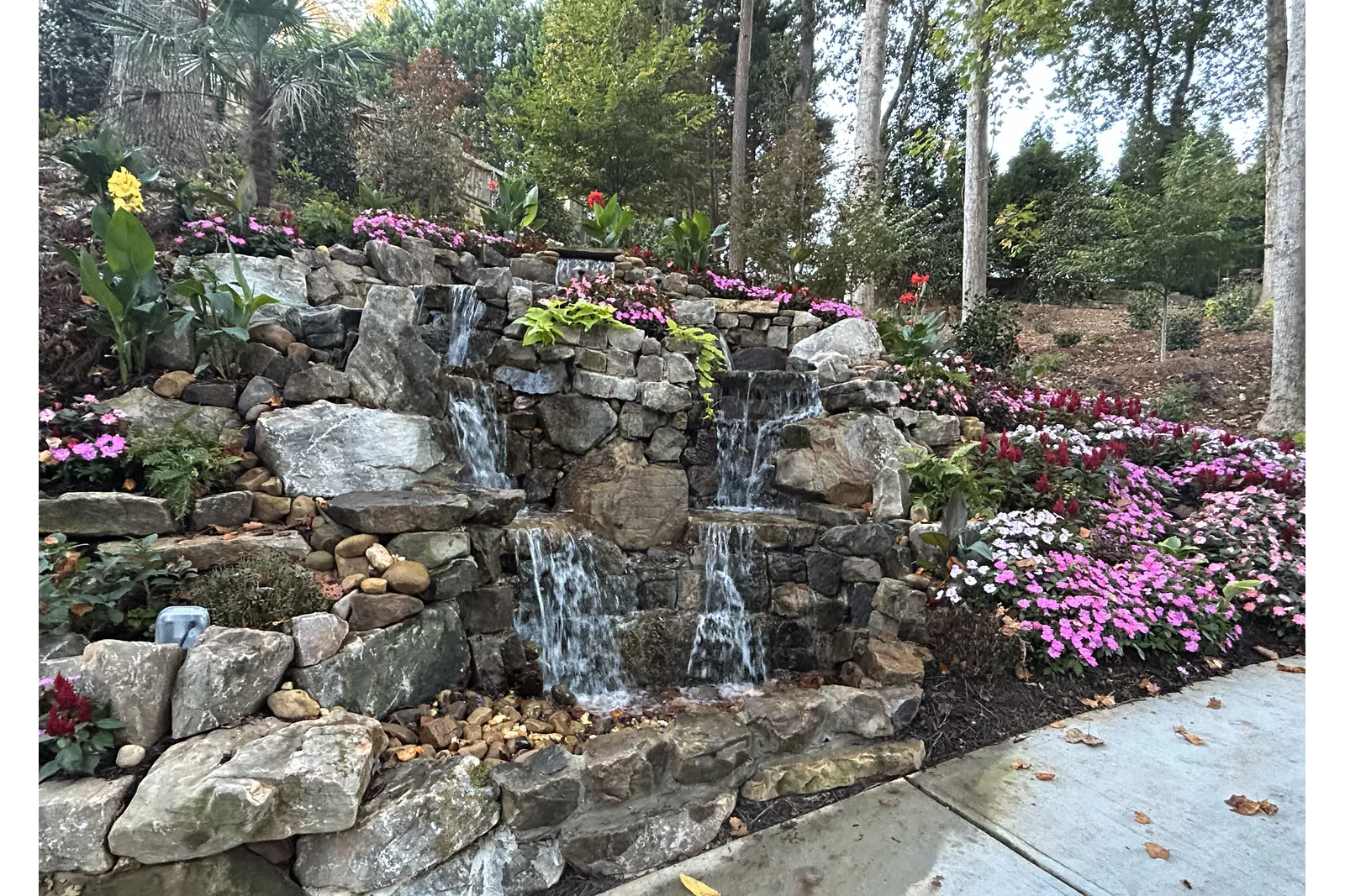 Tiered stone waterfall surrounded by pink and white flowers alongside a wooded garden path on a fall day.