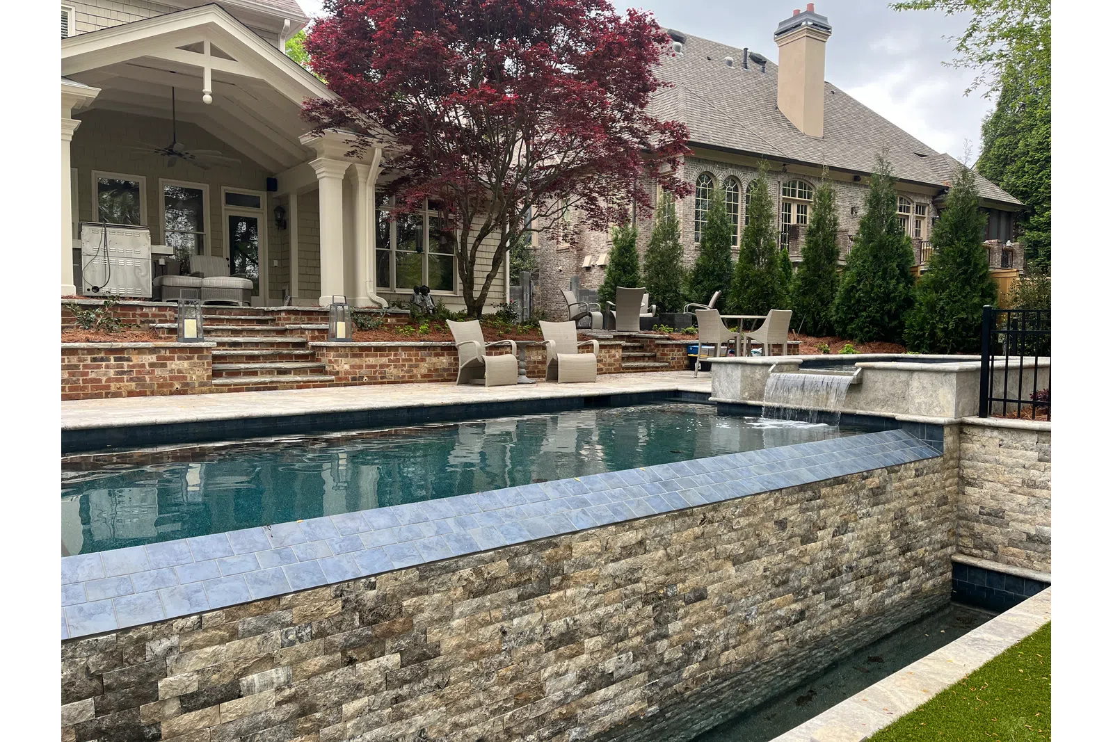 Modern backyard pool with stone walls, waterfall feature, patio seating, and a covered porch beside a large house.