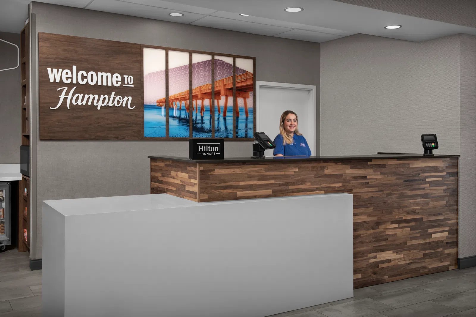 Reception desk at Hampton hotel with a friendly staff member and welcome sign featuring a pier over water.