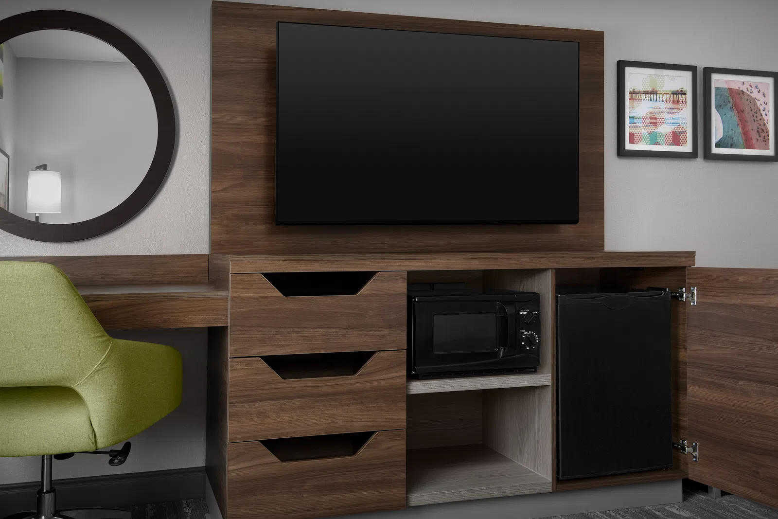 Modern hotel room corner with wood TV stand, mounted flat-screen TV, microwave, mini fridge, green chair, and wall art.