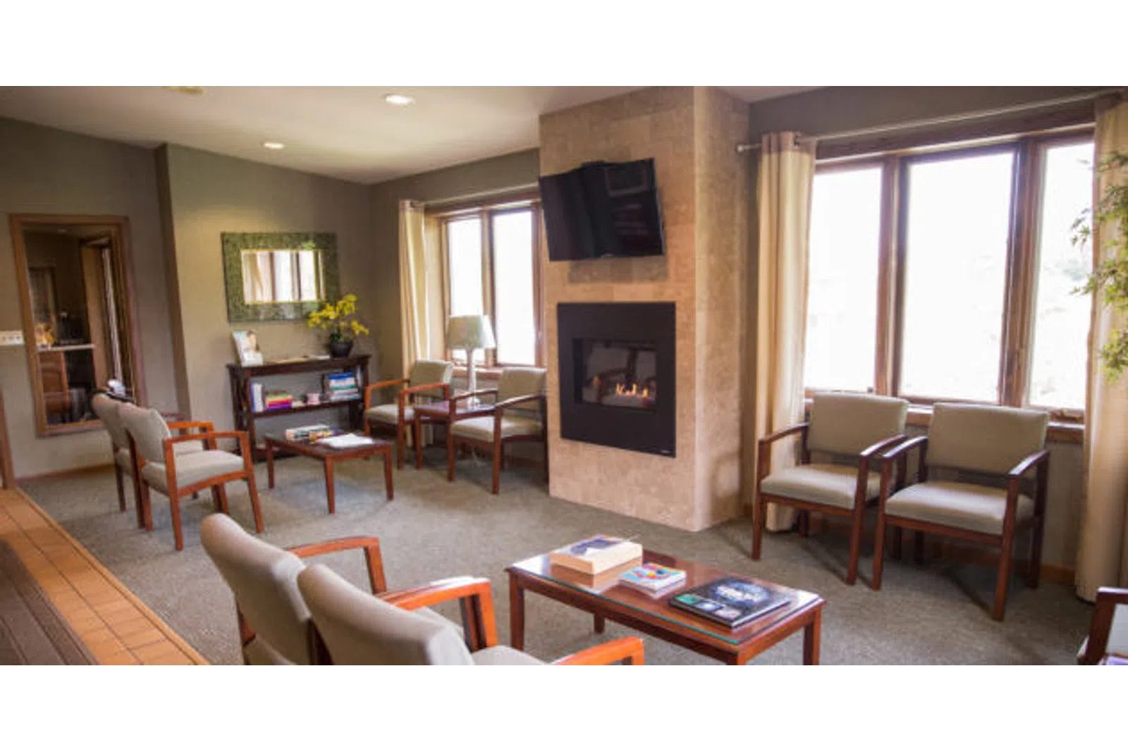 Cozy waiting room with cushioned chairs, a fireplace, flat-screen TV, and natural light from large windows.
